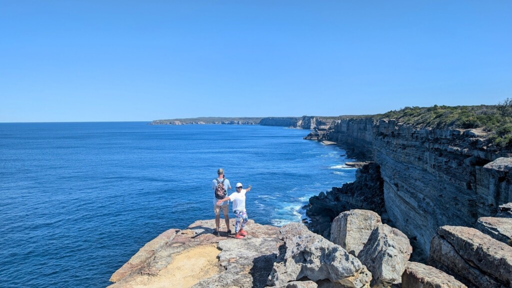 Two people stand on a rocky cliff overlooking the vast blue ocean in Jervis Bay.