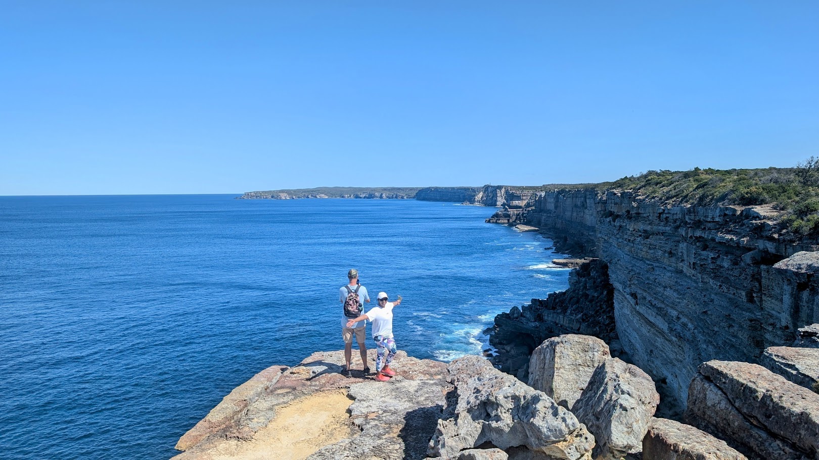 Two people stand on a rocky cliff overlooking the vast blue ocean in Jervis Bay.