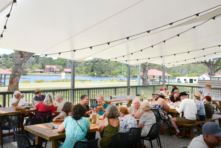 People enjoying drinks and food at outdoor tables under a canopy near a waterway.