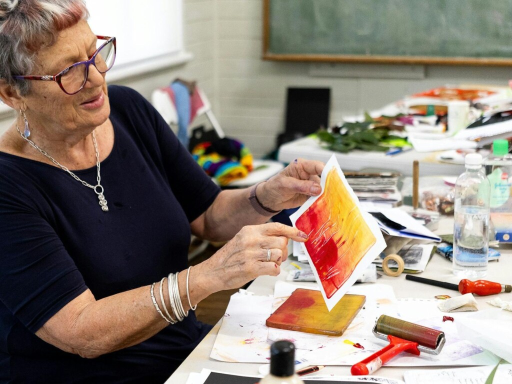 Older woman with purple glasses smiling while holding up a vibrant yellow, orange, and red gel-plate print she created at an Arts Muster workshop in Jervis Bay, with a red ink roller and art supplies on the table.