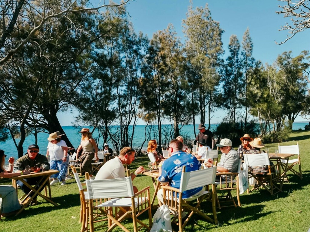 Guests enjoying a 10-course degustation at Coastal Forage Jervis Bay, seated at bamboo tables on a grassy lawn overlooking the turquoise ocean and white sand.