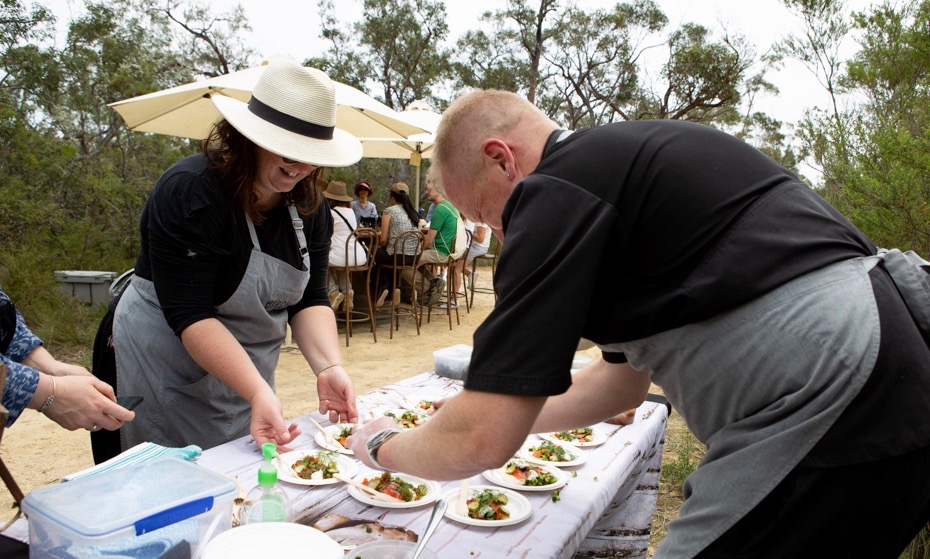 Two chefs in grey aprons carefully plating gourmet appetizers on a white tablecloth at an outdoor dining station in the Australian bush during the Coastal Forage event in Jervis Bay.