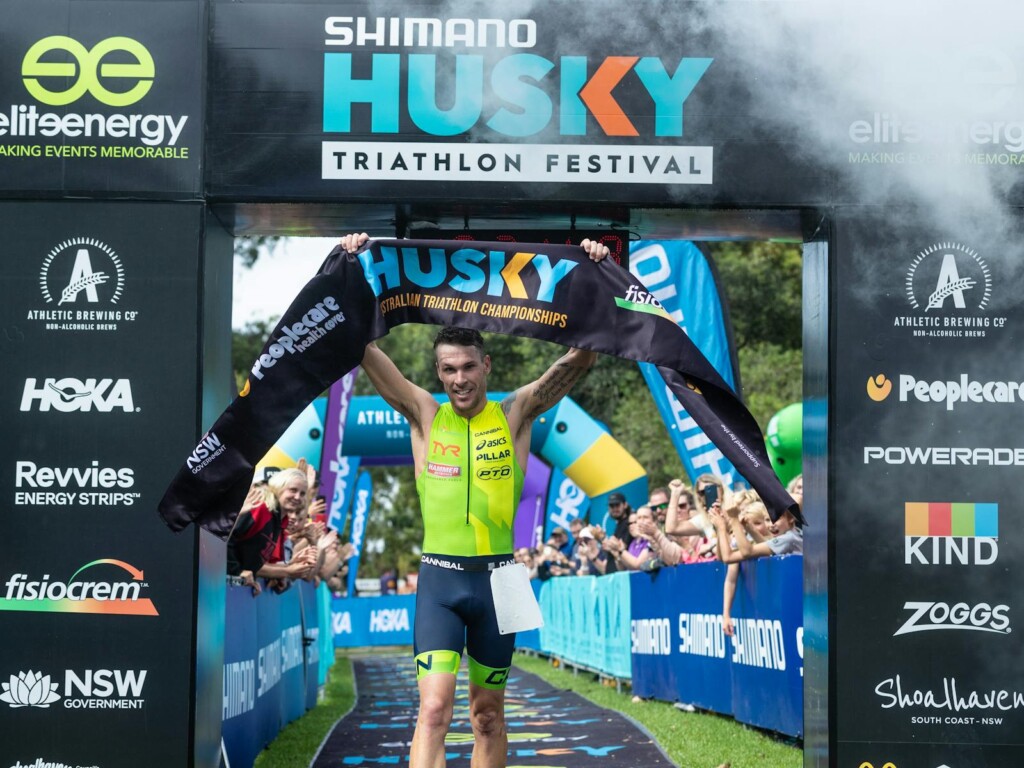 Male triathlete in a lime green suit crossing the finish line of the Shimano Husky Triathlon Festival in Jervis Bay with a celebratory fist pump.