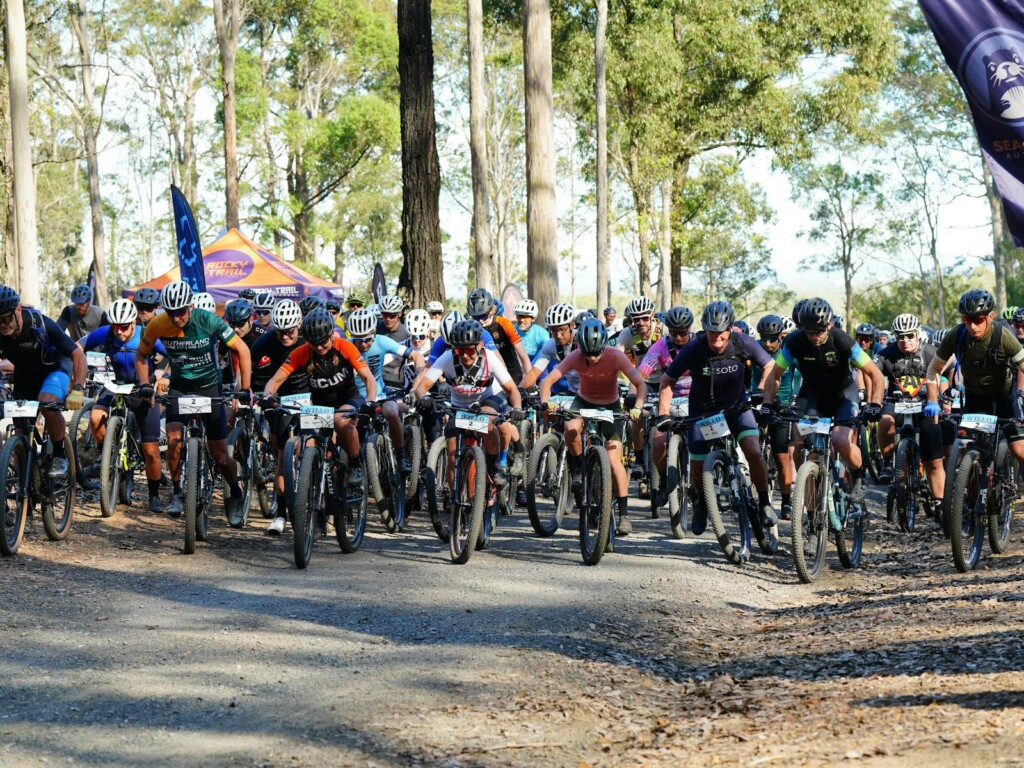 Mountain bikers lined up at the vibrant start line of The Willo Mountain Bike Marathon on a dirt track surrounded by tall eucalyptus trees, hosted by Rocky Trail Entertainment.
