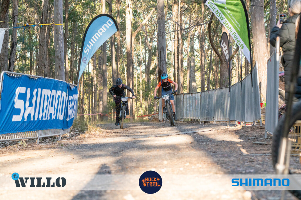 Two young mountain bikers racing side-by-side on a dirt trail lined with metal barriers and blue Shimano event banners in a sunny forest.