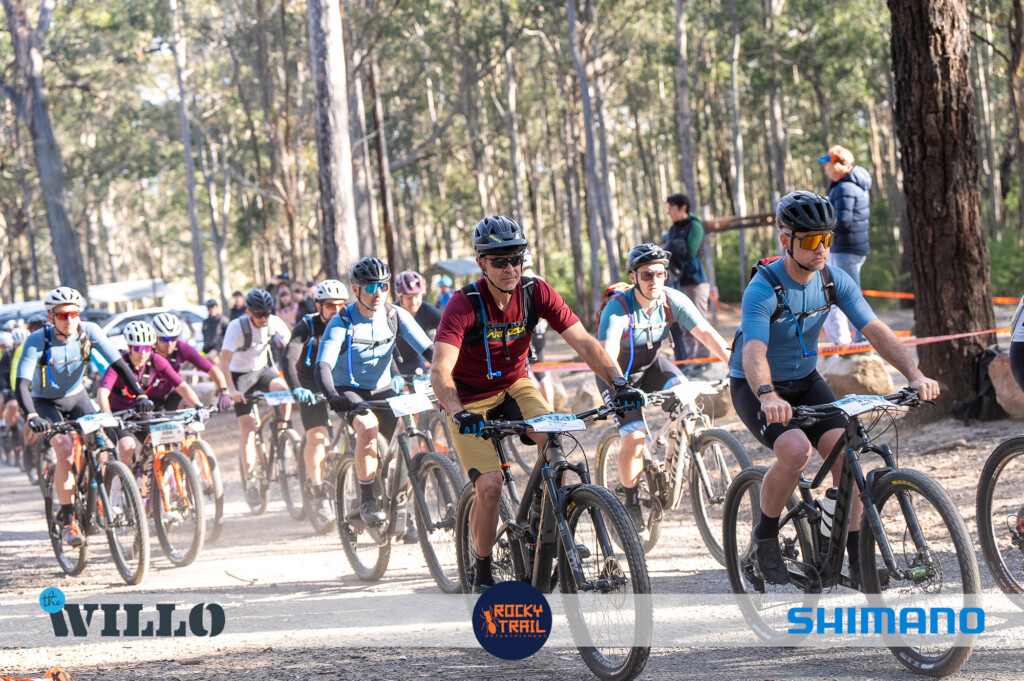 A large group of adult mountain bikers riding together on a wide dirt trail through a eucalyptus forest, wearing helmets, hydration packs, and racing numbers at The Willo event.