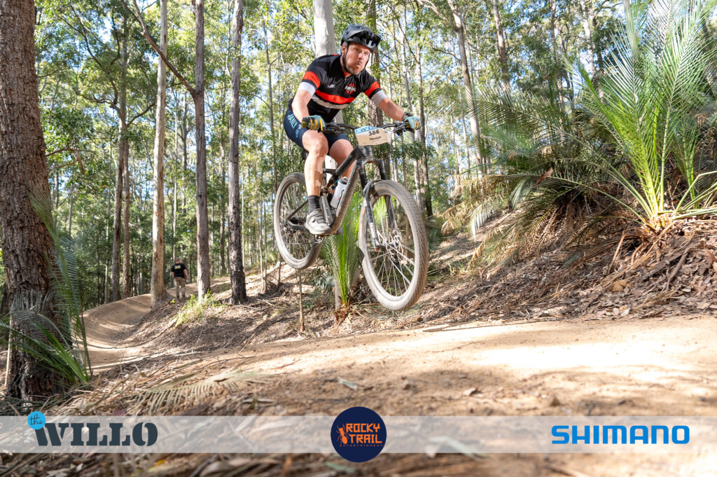 A male mountain biker in a black and red jersey catching air over a dirt jump on a forest trail surrounded by green ferns and tall trees during The Willo mountain bike race.