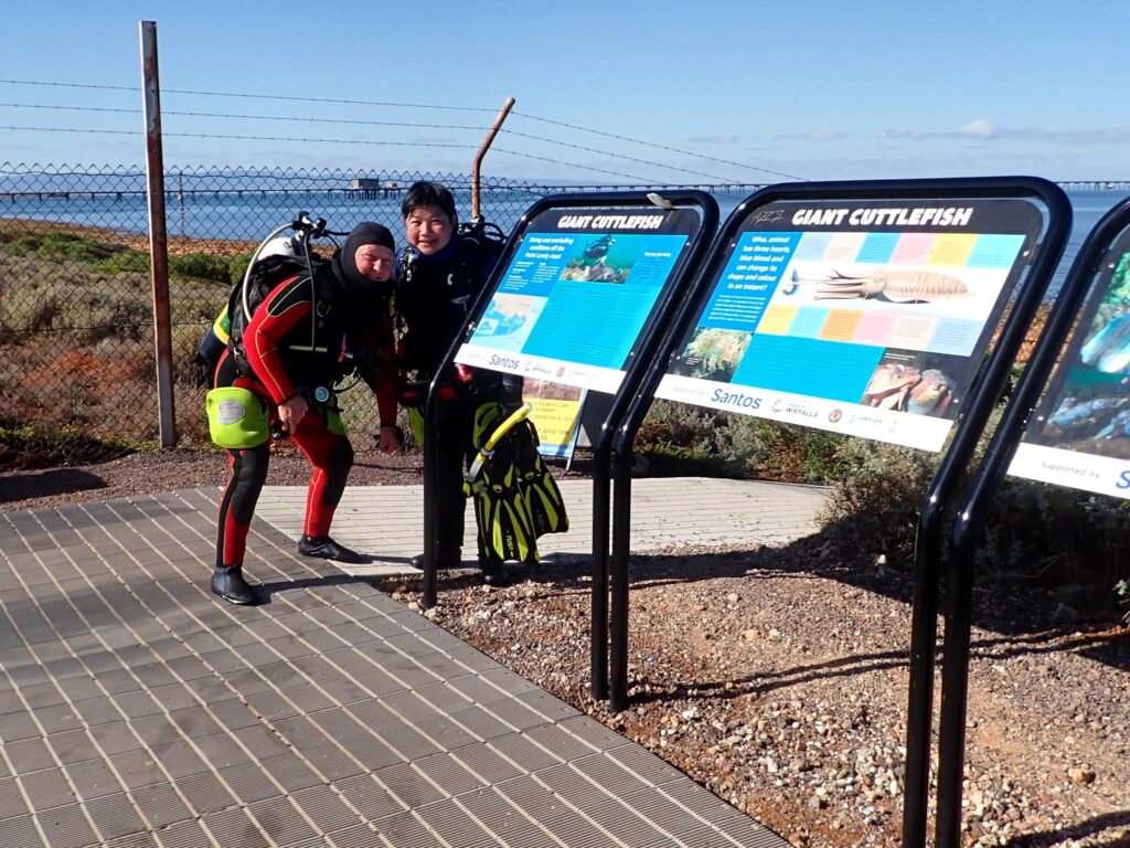 Two fully geared scuba divers smiling next to Giant Cuttlefish informational signs before entering the water.