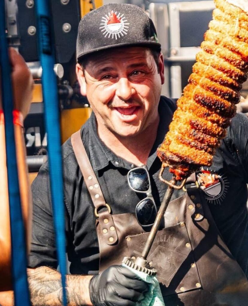 A cheerful pitmaster in a leather apron proudly holding a massive skewer of crispy, slow-roasted meat at the Kangaroo Valley BBQ Festival.