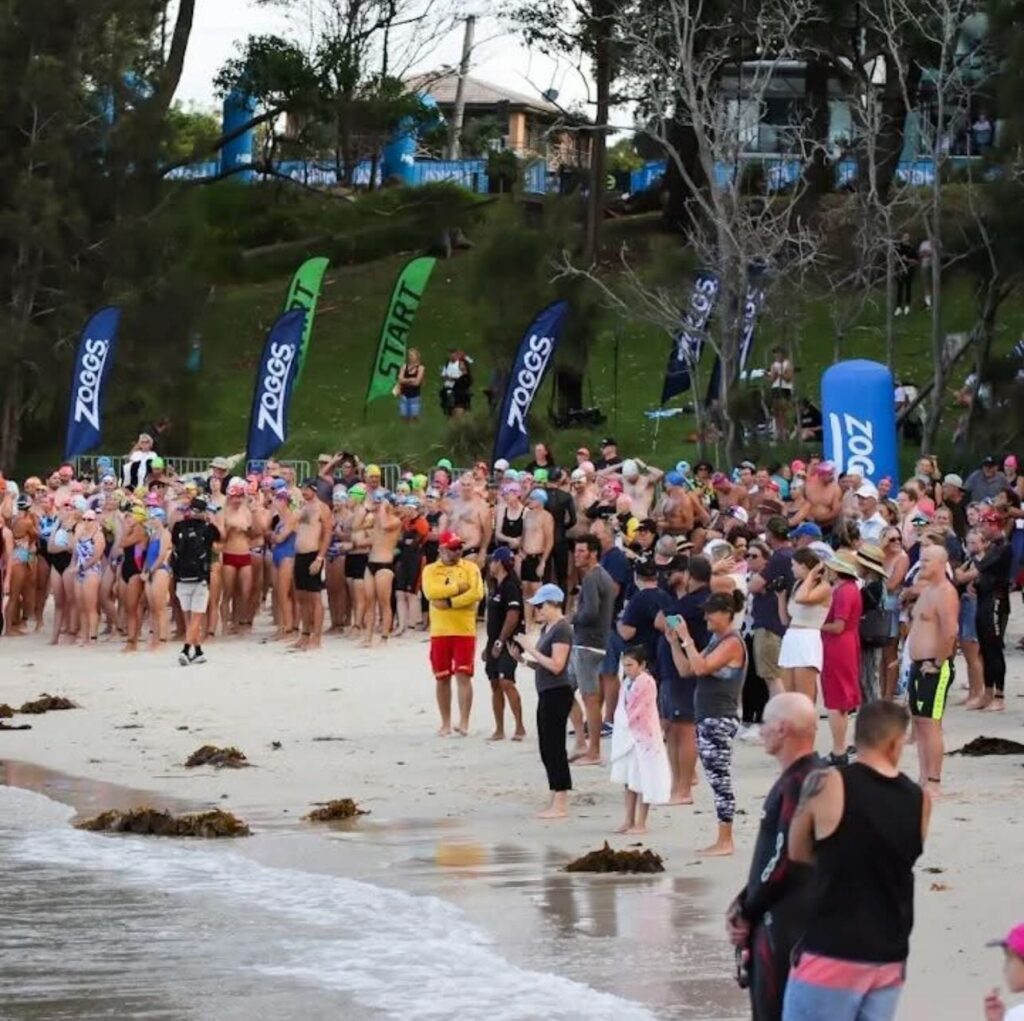 Hundreds of eager triathletes wearing colorful swim caps gather on the white sands of Jervis Bay for the swim start of the Shimano Husky Triathlon Festival.