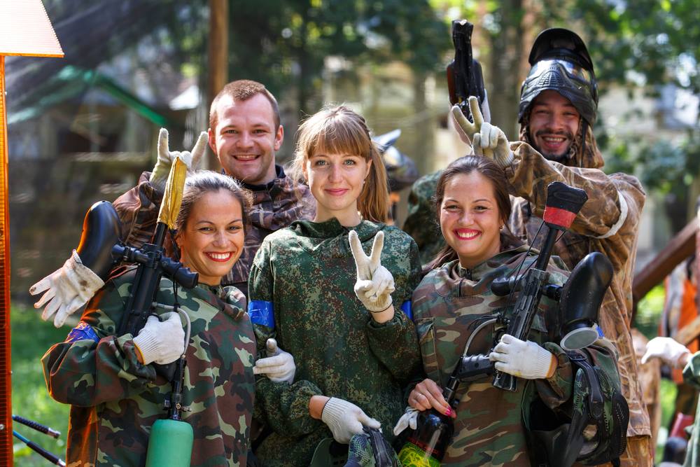 A smiling group portrait of five friends (3 women, 2 men) in assorted military-style camouflage gear and safety masks, holding paintball markers after a game at Jervis Bay. One woman makes a peace sign. Natural bush lighting.