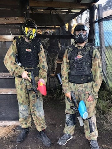 A vertical portrait photo of two young men in full camouflage suits, tactical vests, and protective masks (one yellow, one black) standing in the dirt staging area of Shoalhaven Skirmish, holding markers with red and blue hoppers. Background shows other players.