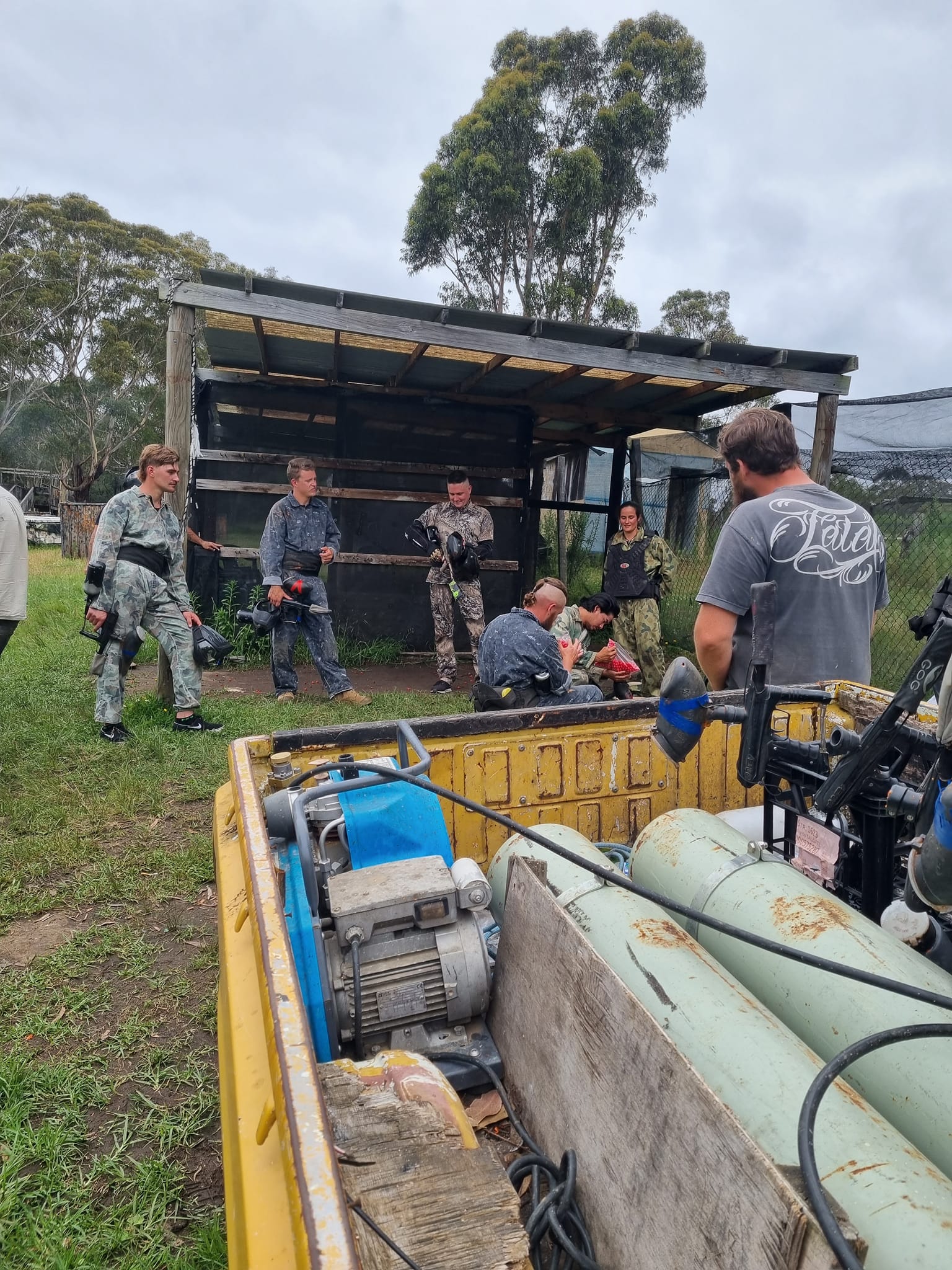 The rear of a yellow utility truck holding large green gas tanks and a gas compressor/motor unit in the foreground, with an overcast sky. In the background, a supervised staging hut is visible, where several players in camouflage gear are interacting.
