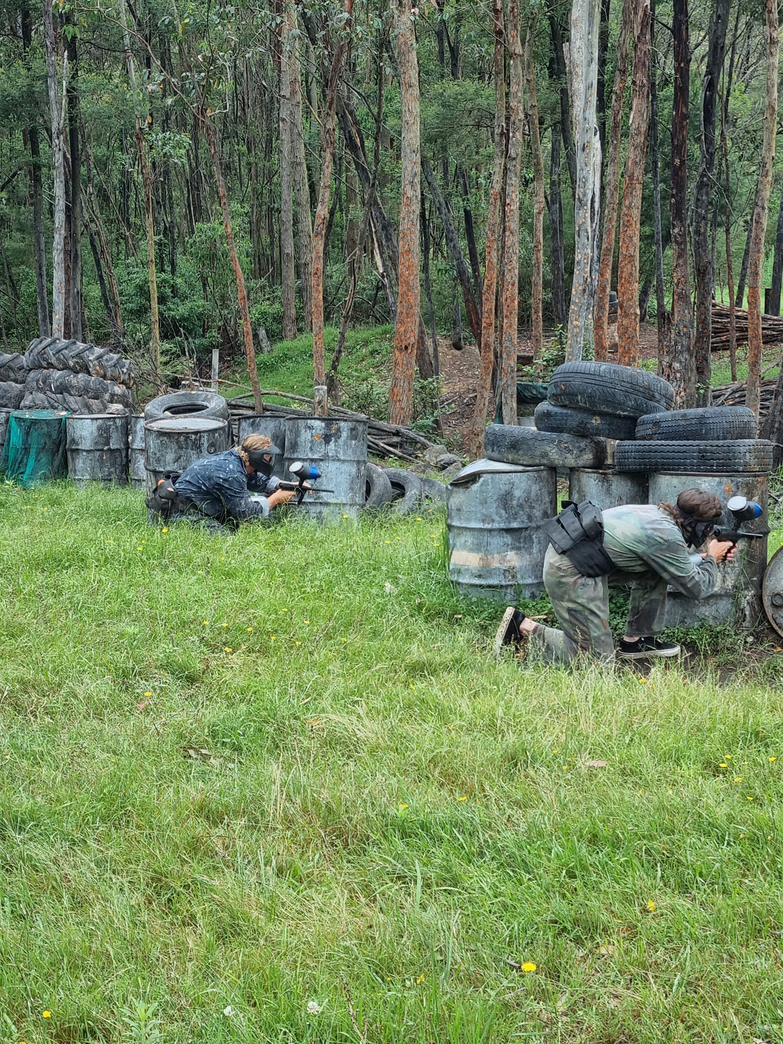 Close-up action shot of two paintball players in tactical camouflage gear and full protective masks taking cover behind a white industrial container (IBC tank) at Shoalhaven Skirmish Jervis Bay. One player wears a high-visibility orange vest over camo in bright sunlight, aiming a marker with a prominent red hopper.