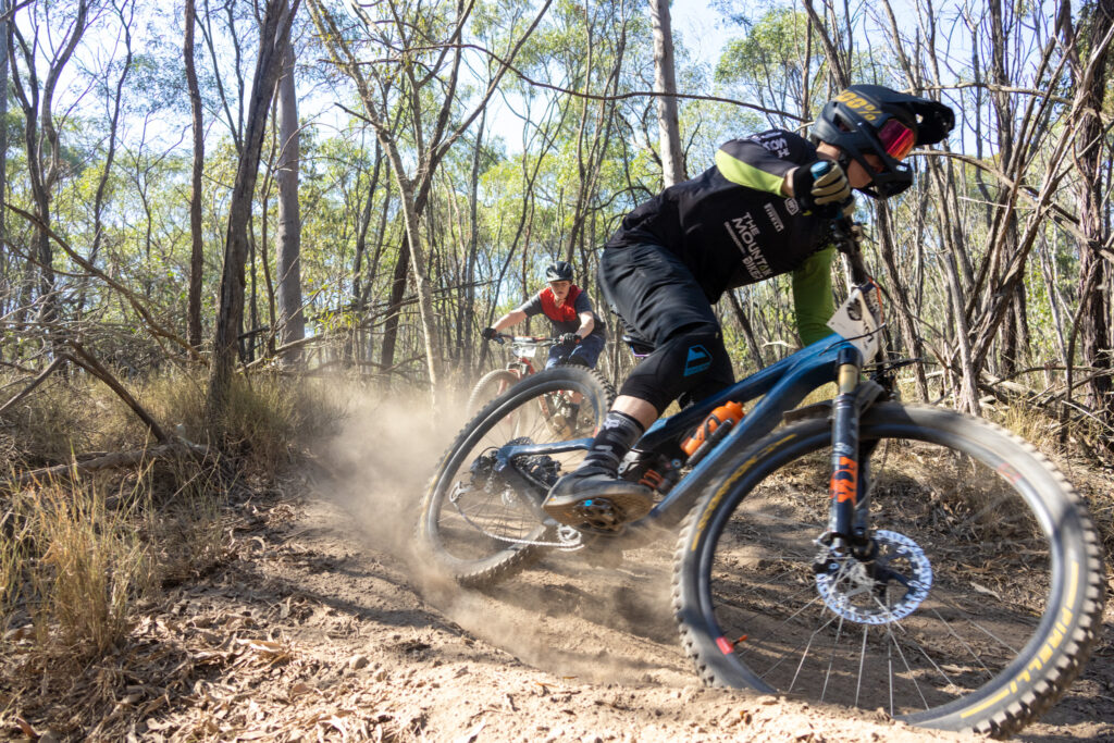 A mountain biker leaning hard into a dusty corner on a dry dirt trail, kicking up a cloud of dust in a sparse eucalyptus forest.