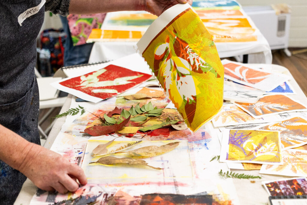 A person's hands holding up a freshly printed botanical artwork in vibrant yellow and green, with real eucalyptus leaves and ferns scattered on a printmaking table at the Jervis Bay Arts Muster.