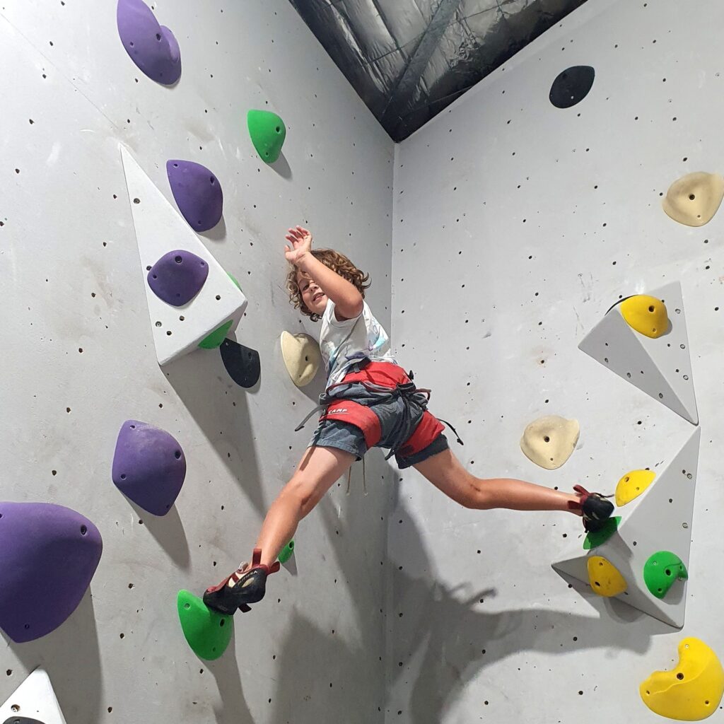 A young boy with curly hair joyfully bouldering at Indoor Climb South Coast, stretching between vibrant green and yellow holds on a modern geometric climbing wall.