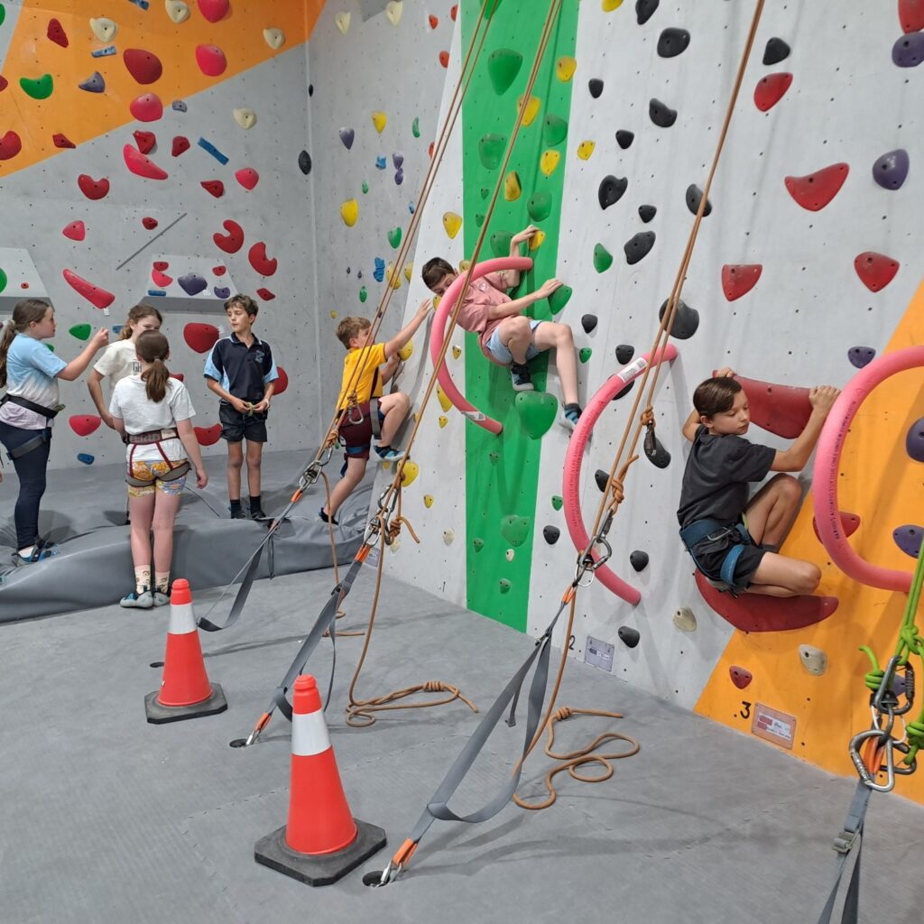 A group of children participating in a climbing class at Indoor Climb South Coast, using pink hula hoops as training tools on a green and grey top-rope wall.