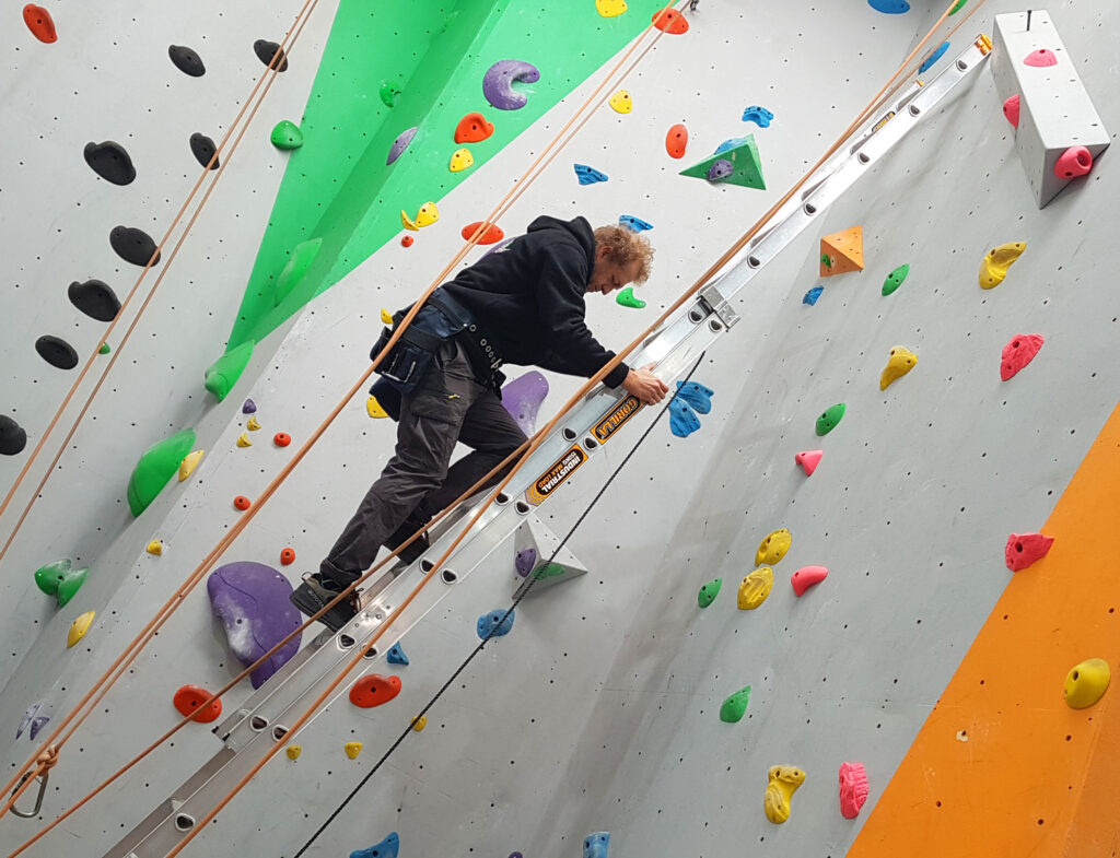 A professional route setter at Indoor Climb South Coast using an industrial ladder to meticulously place new colorful climbing holds on a tall indoor wall.