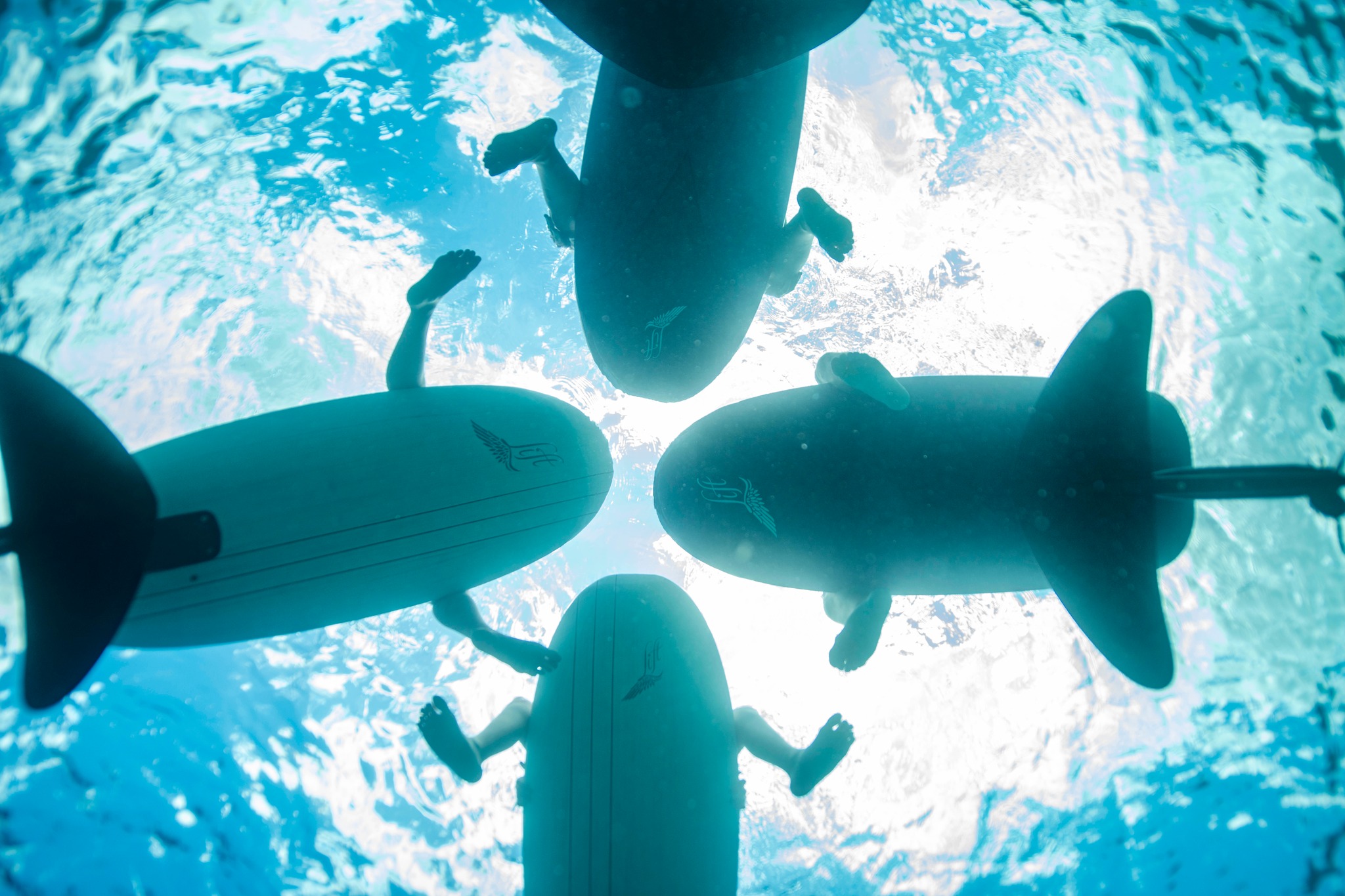 Underwater perspective looking up at the bottoms of four eFoil boards arranged in a circle, with riders' legs dangling in the sunlit, turquoise water of Jervis Bay.