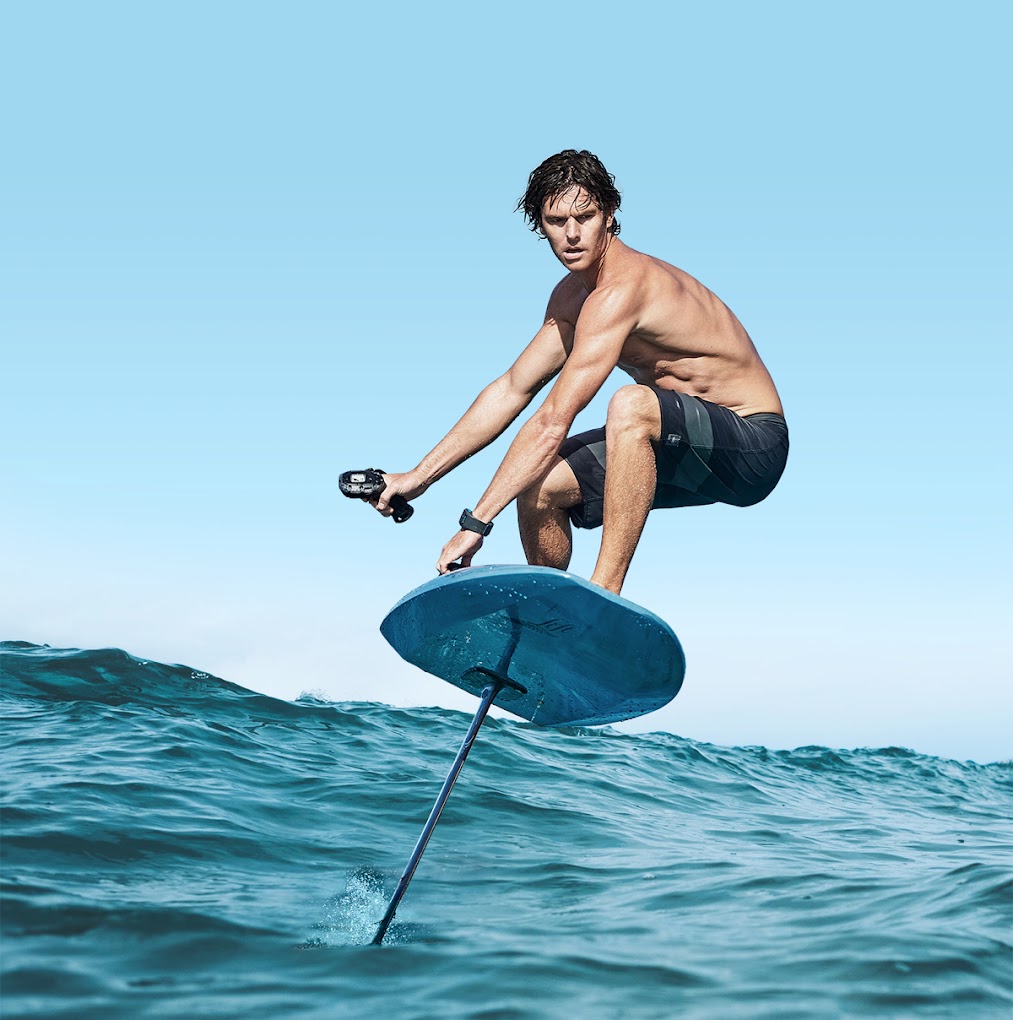 A fit, shirtless man riding a blue electric hydrofoil board (eFoil) high above the turquoise waters of Jervis Bay under a clear blue sky, holding a wireless controller.