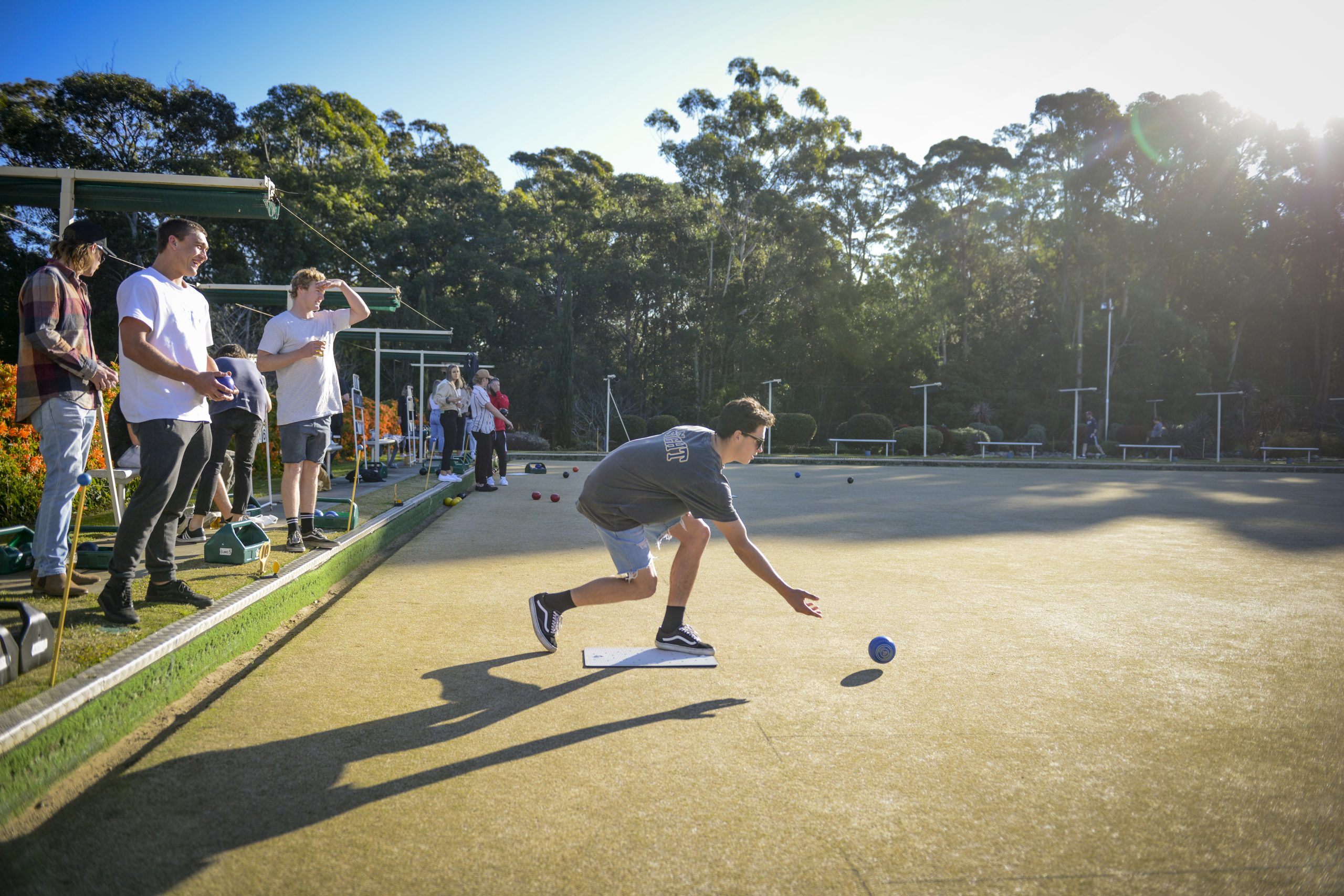 A young man in sunglasses bowling a blue ball on a sun-drenched lawn bowls green at Husky Sports, Jervis Bay, with friends watching in the background.