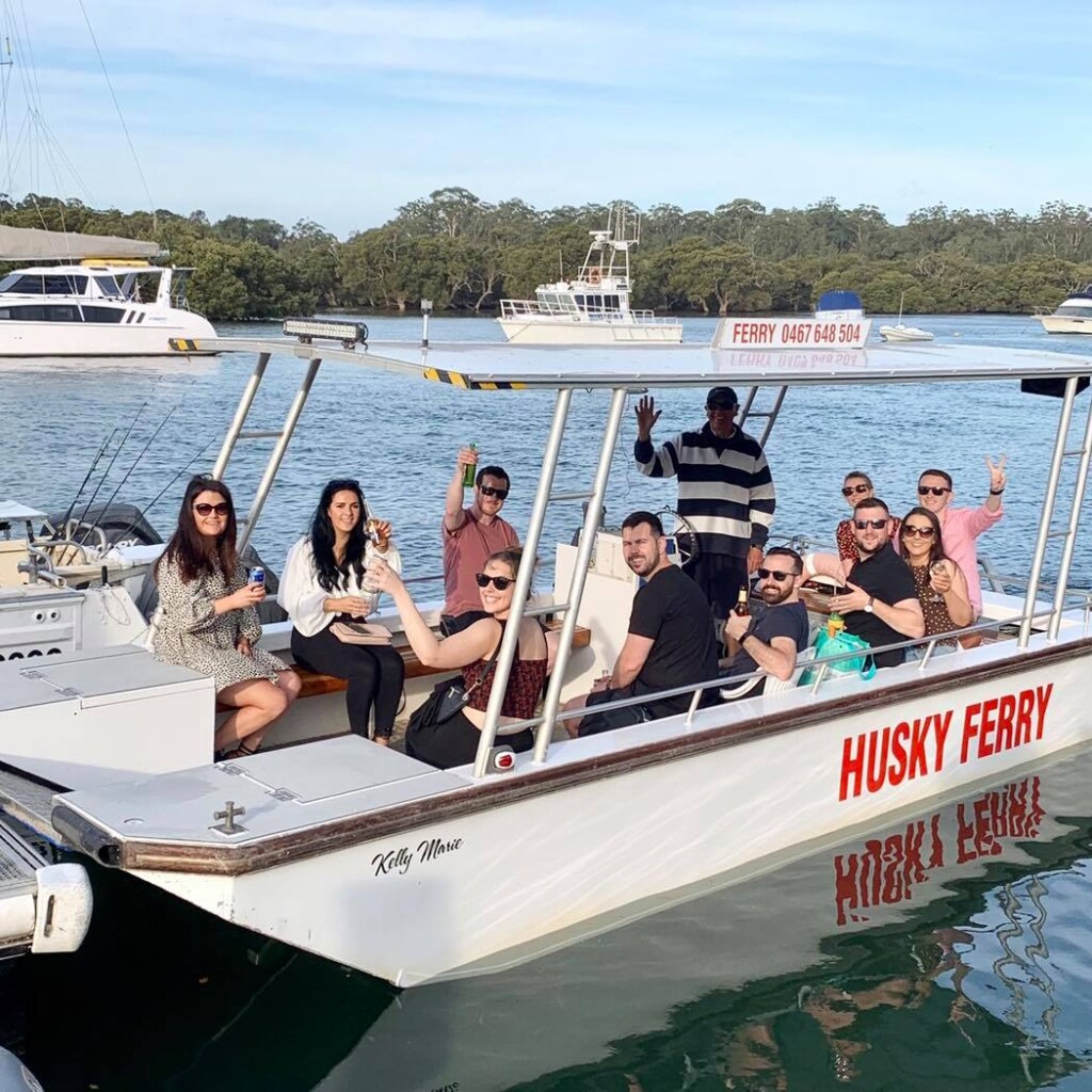 A group of seven friends smiling and raising their drinks in a toast while enjoying a sunny day cruise on the Husky Ferry on Currambene Creek, Jervis Bay, with the captain waving from the helm.
