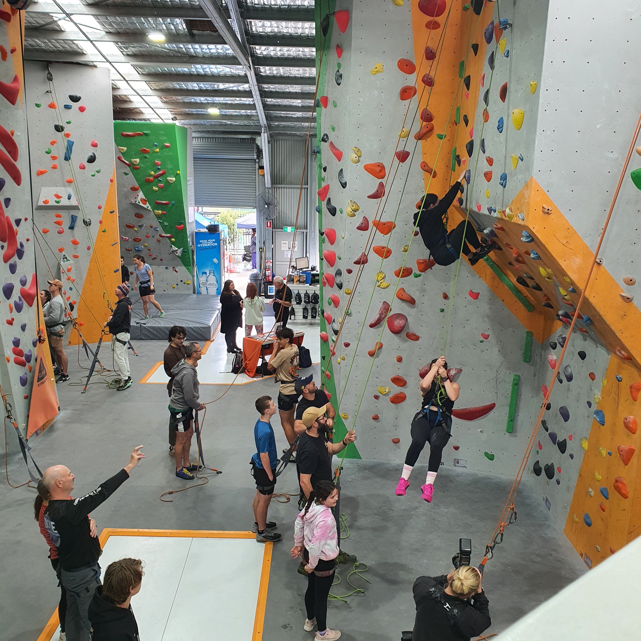 A vibrant indoor rock climbing gym at Indoor Climb South Coast featuring towering walls with colorful holds, climbers scaling routes, and supportive instructors assisting beginners on safety mats under a bright skylit warehouse roof.