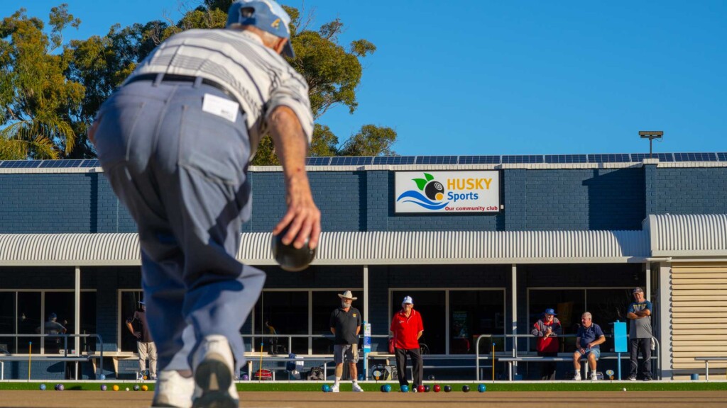 A vibrant group of people playing barefoot lawn bowls on a sunny afternoon at Huskisson Sports Club in Jervis Bay, surrounded by green trees.