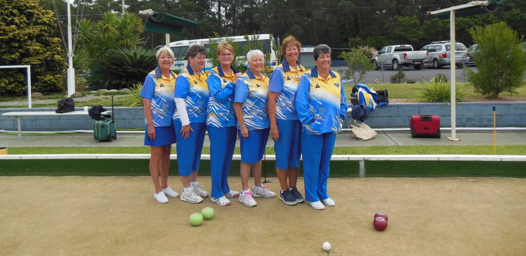 Six smiling women from the Huskisson lawn bowls team wearing matching blue, yellow, and white uniforms, standing together on the green.