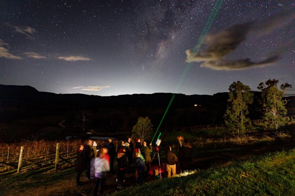 A group of people sitting in camping chairs on a dark, sandy beach in Jervis Bay, gazing up at a bright, moonlit sky with scattered clouds, illuminated by the red safety light of a nearby telescope.