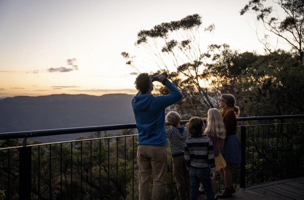 A family of five standing on a lookout platform surrounded by eucalyptus trees, gazing out over distant layered mountains under a warm, golden sunset sky, with the father looking through binoculars.