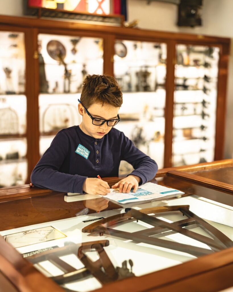 A young boy wearing glasses fills out his Jervis Bay Maritime Museum Treasure Hunt booklet on top of a glass display case featuring historic wooden navigational instruments.
