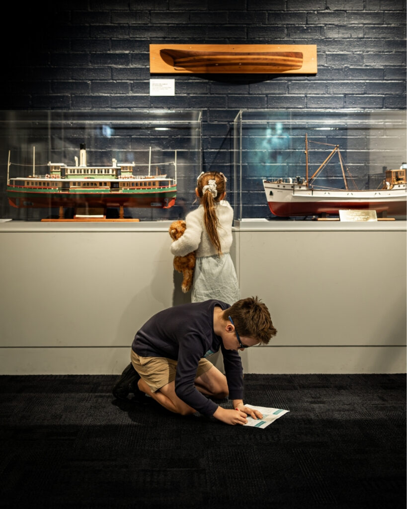 Two children exploring the Jervis Bay Maritime Museum, with a boy kneeling on the floor to write on his treasure hunt map while a girl admires detailed model ships.