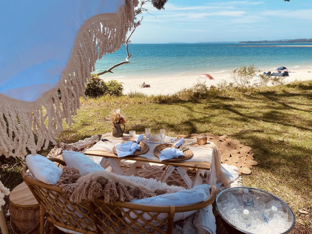 Luxury boho picnic setup on a grassy foreshore overlooking the pristine white sand and turquoise water of Jervis Bay.