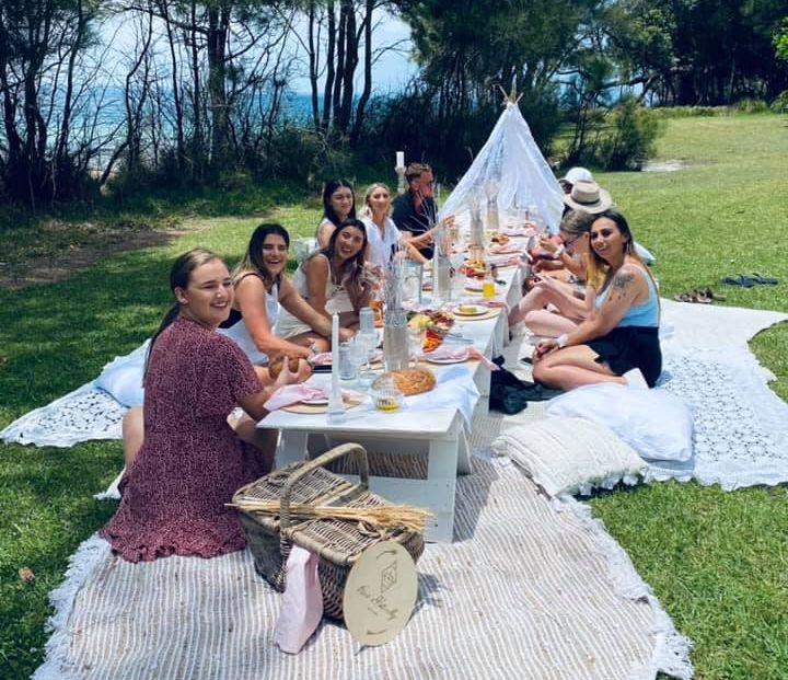 A luxury bohemian styled picnic setup on the white sand of Jervis Bay featuring a low wooden table, grazing board, velvet cushions, and a fringe umbrella at sunset.