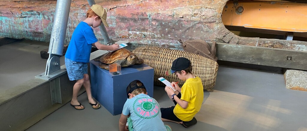 Two children smiling and reading a treasure hunt booklet in front of the historic Lady Denman wooden ferry at the Jervis Bay Maritime Museum in Huskisson.