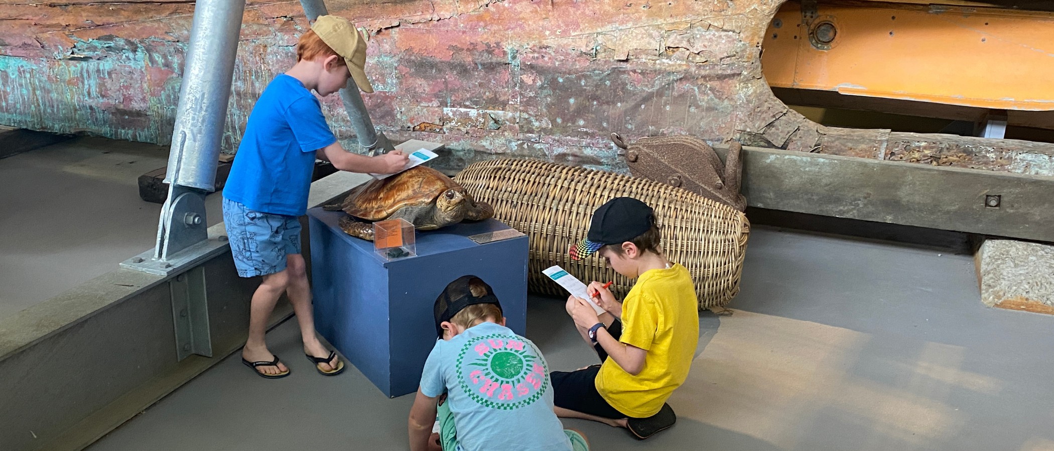 Two children smiling and reading a treasure hunt booklet in front of the historic Lady Denman wooden ferry at the Jervis Bay Maritime Museum in Huskisson.
