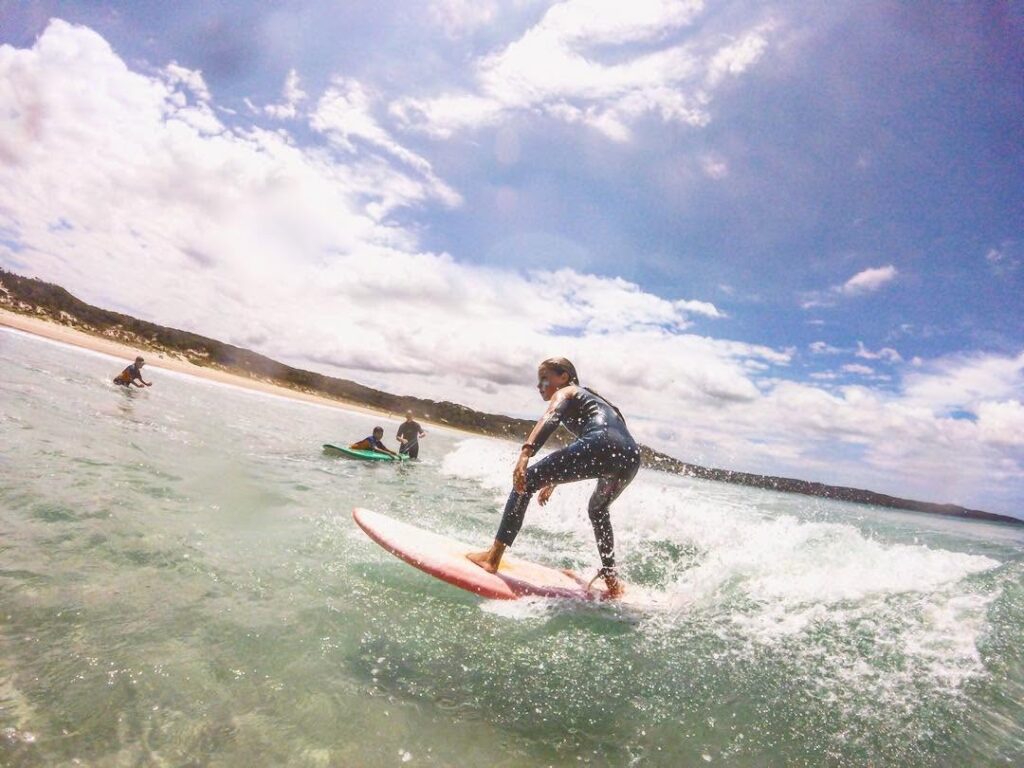 A surfer in a wetsuit riding a soft-top board on a gentle wave at a Jervis Bay beach with other surfers nearby.
