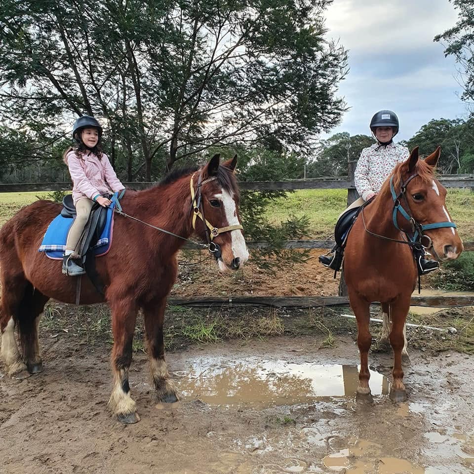 Two young girls in helmets and riding jackets sit atop horses in a paddock, facing the camera near a large, muddy puddle, with native Australian trees and fencing visible in the background.