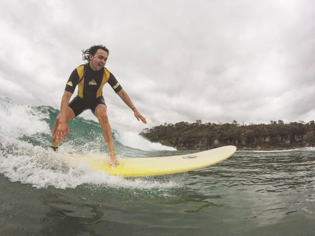 A young man smiling while surfing on a yellow longboard at a beach with rocky cliffs in the background under an overcast sky.
