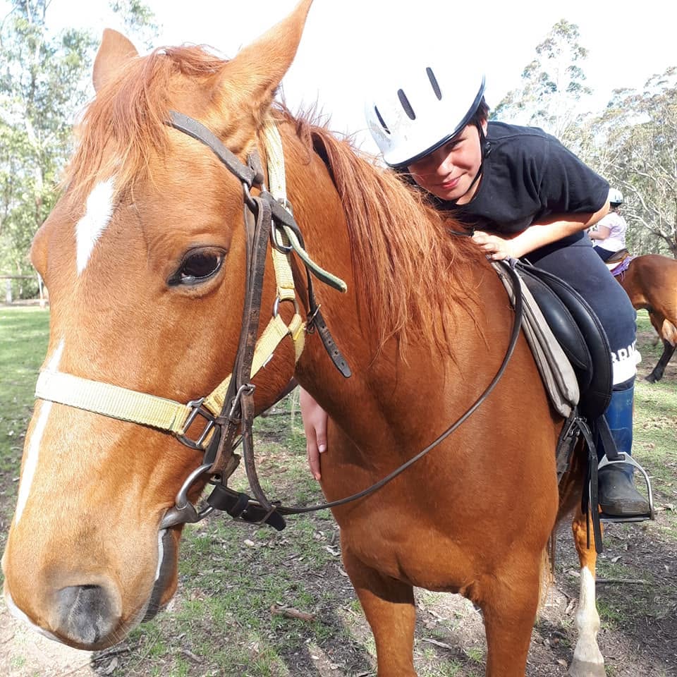 A close-up portrait of a young child wearing a white riding helmet, smiling while leaning over the neck and back of a chestnut horse.