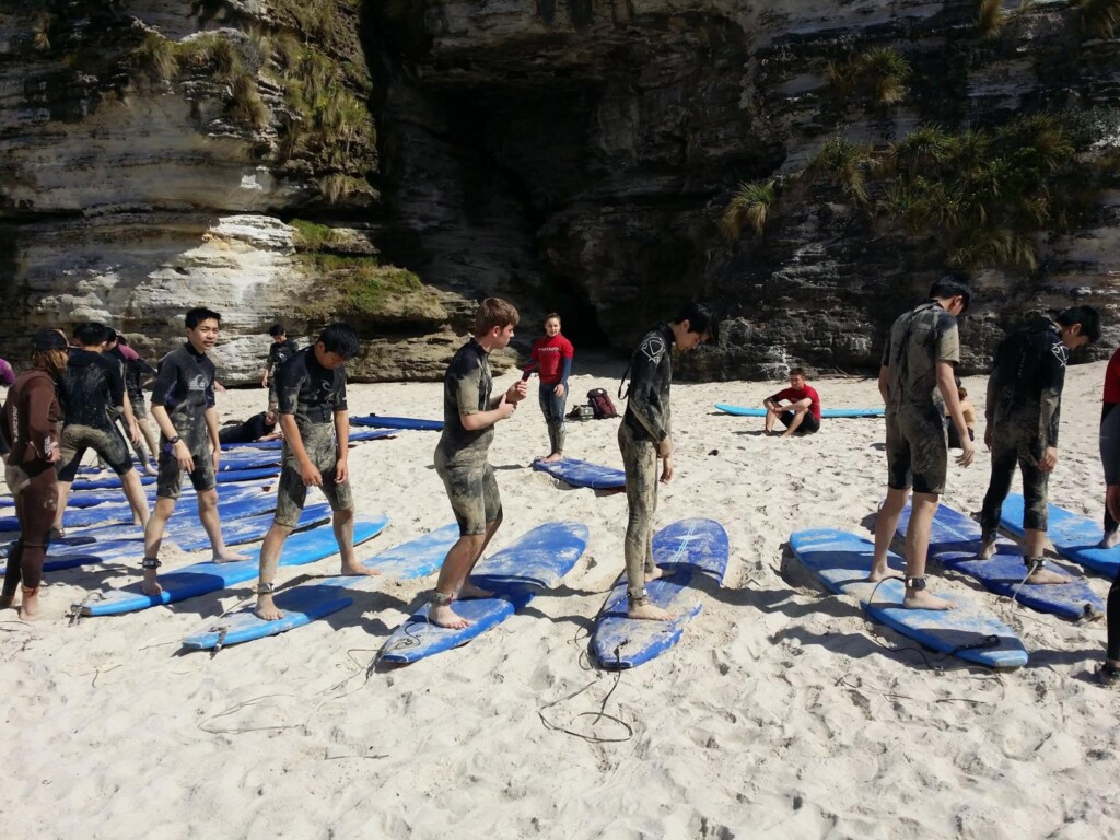 A group of surf students practicing their pop-up technique on boards laid out on the white sand of a beach with rock faces nearby.