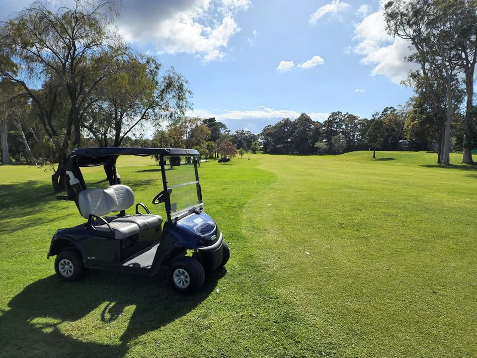 A black golf cart parked on the edge of a lush green fairway at St Georges Basin Golf Club, with a clear sky and scattered white clouds overhead.