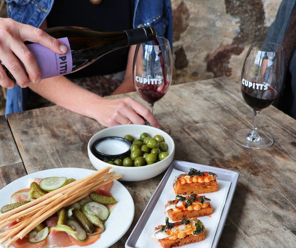 Close-up of a person pouring Cupitt's Estate red wine at a rustic timber table with green olives, cured meats, and anchovy toast.