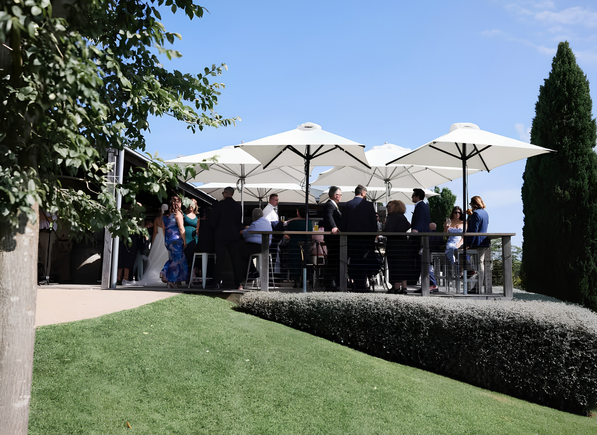 Guests networking at an outdoor corporate event on a landscaped terrace with white umbrellas under a clear blue sky.
