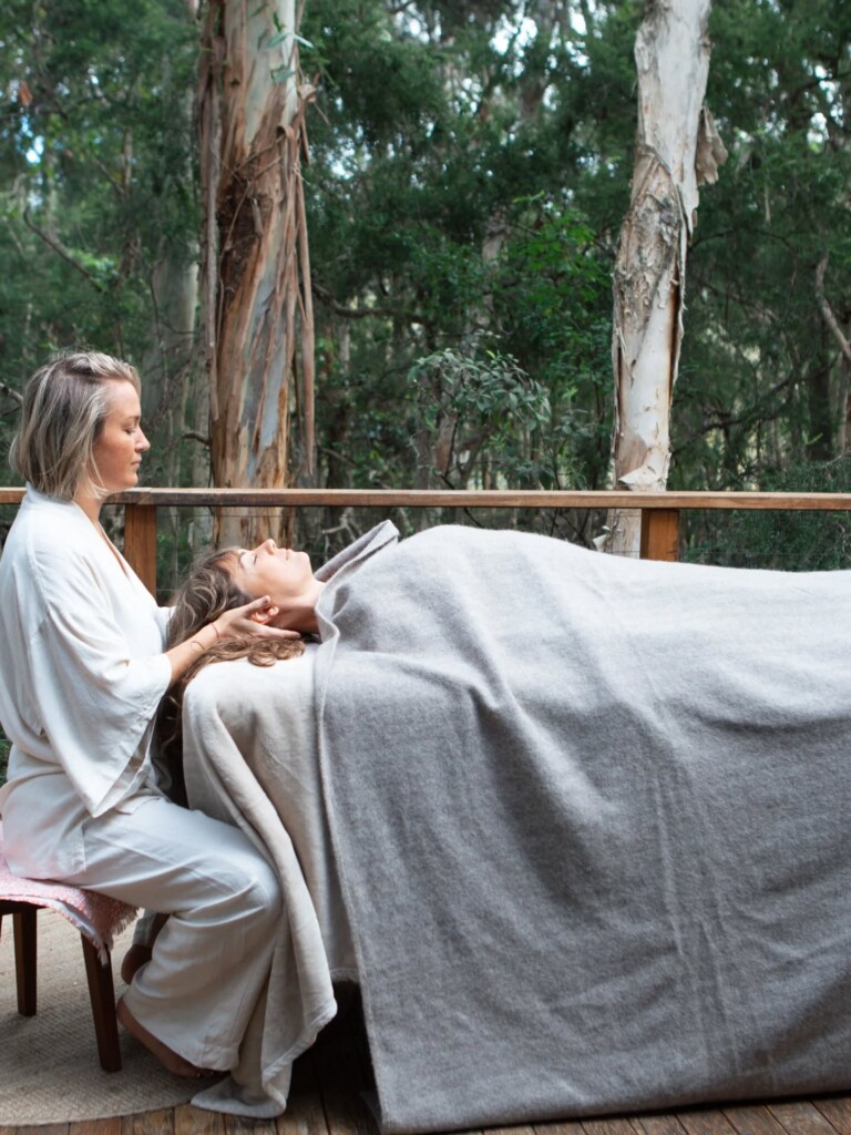 A guest receiving a relaxed head massage on a massage table set up on a wooden verandah surrounded by trees.