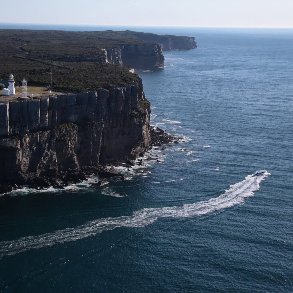 Aerial view of a white tour boat leaving a wake in dark blue water as it cruises past the towering, layered cliffs and lighthouse of Point Perpendicular.