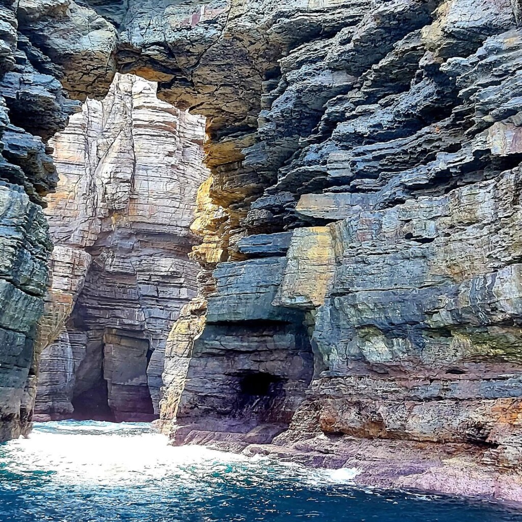 Close-up of heavily fractured, grey and brown layered sandstone cliff face featuring a natural rock arch over churning blue water.