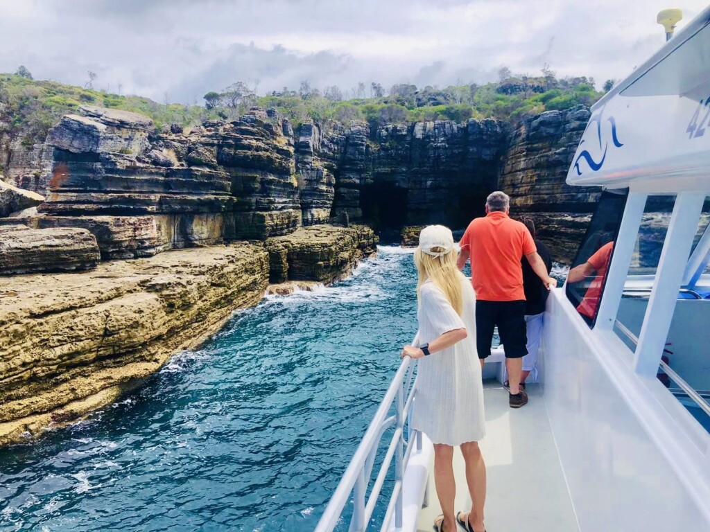 Passengers on the deck of a white tour boat looking out at a dark sea cave cut into stepped, layered sandstone cliffs.